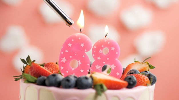 흰 구름과 분홍색 하늘을 배경으로 딸기와 라이터가 있는 아름다운 케이크에 생일 케이크 번호 68 분홍색 촛불이 있습니다.