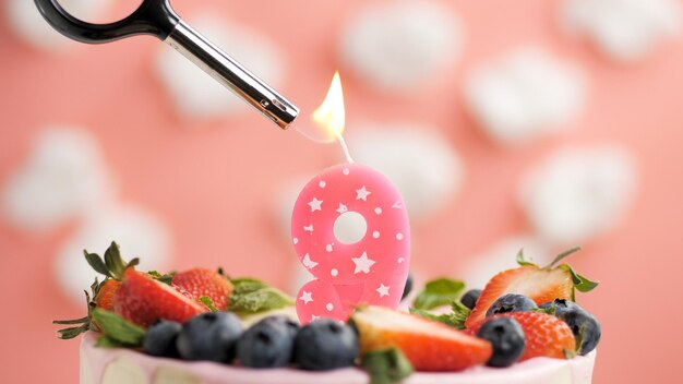 Birthday cake number 9 pink candle on beautiful cake with\
berries and lighter with fire against background of white clouds\
and pink sky closeup view