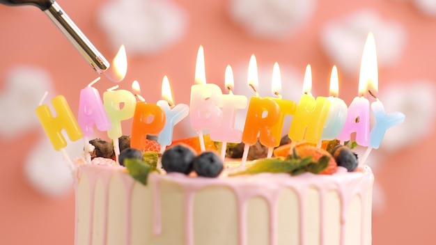 Birthday cake with candle title Happy Birthday on beautiful cake with berries and lighter with fire against background of white clouds and pink sky Closeup view