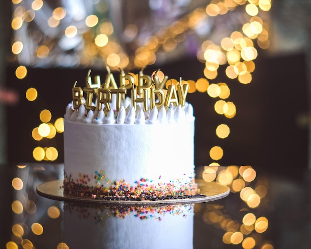 Birthday cake with candles garland with bright bokeh lights on\
the background the white cake is decorated with colored sprinkles\
and stands on a reflective surface