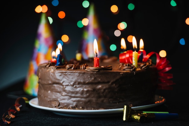 Birthday cake with an illuminated candle against light backdrop and party hat