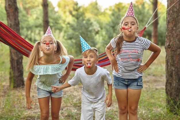 誕生日、子供時代、お祝いのコンセプト-パーティーの角を吹いて夏の屋外で楽しんでいる幸せな子供たちのクローズアップ