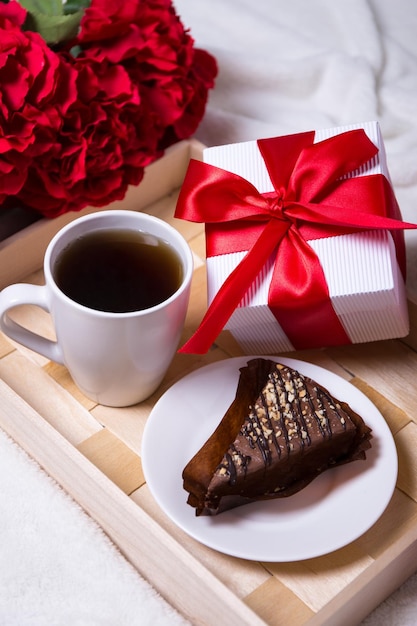 Birthday concept - close up of breakfast with chocolate cake, tea and little gift on wooden tray and red flowers