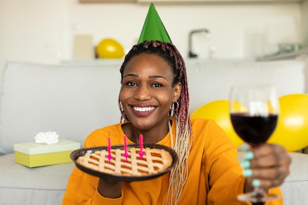 Birthday concept excited black lady holding glass of wine and pie with candles looking and smiling to camera