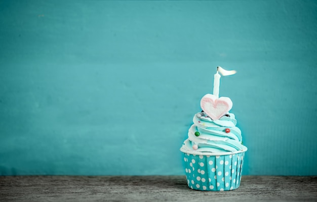 birthday cupcake with sweet heart shape of marshmallow and candle against blue background