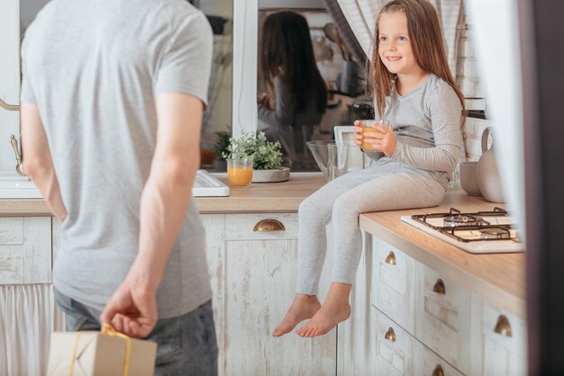 Birthday gift Little girl sitting in kitchen with glass of juice smiling expecting surprise present from her loving father