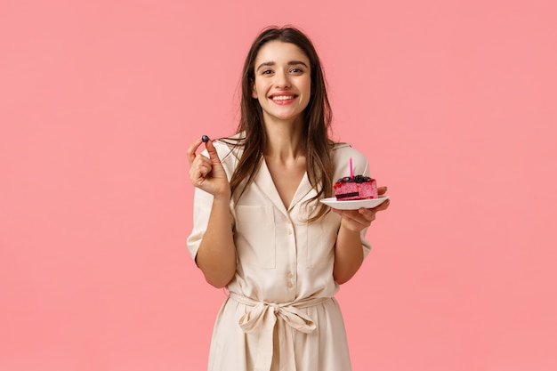 Birthday girl eating delicious cake, blowing-out candle and smiling joyfully, holding blueberry, having party celebrating b-day with friends, grinning standing in dress over pink wall