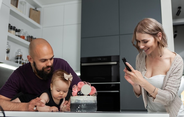 Photo birthday happy family concept young mother takes pictures of her daughter and husband with a cake in kitchen