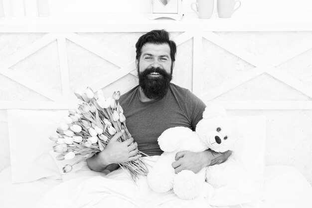 Birthday holiday Fresh flowers Bearded hipster in bed Valentines day gift Man hold bouquet relaxing in bed Flowers delivery service Make surprise concept Gift for spouse Cute teddy bear