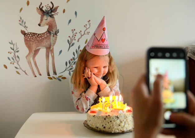 Compleanno di una bambina, seduta vicino al tavolo, guardando una torta con le candele, in sfocatura in primo piano viene scattata una foto al telefono.