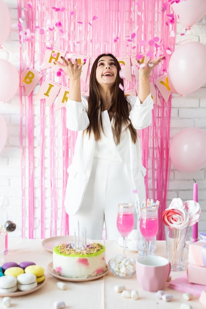 Birthday party birthday tables attractive brunette woman in
white party clothes preparing birthday table with cakes cakepops
macarons and other sweets throwing pink confetti