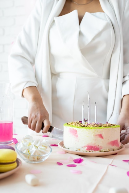 写真 誕生日パーティー誕生日テーブル白いパーティー服を着た魅力的な女性がケーキケーキポップマカロンや他のお菓子で誕生日テーブルを準備してケーキを切る