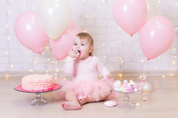 Concetto di festa di compleanno - bambina divertente che mangia la torta sopra il fondo del muro di mattoni con luci e palloncini colorati