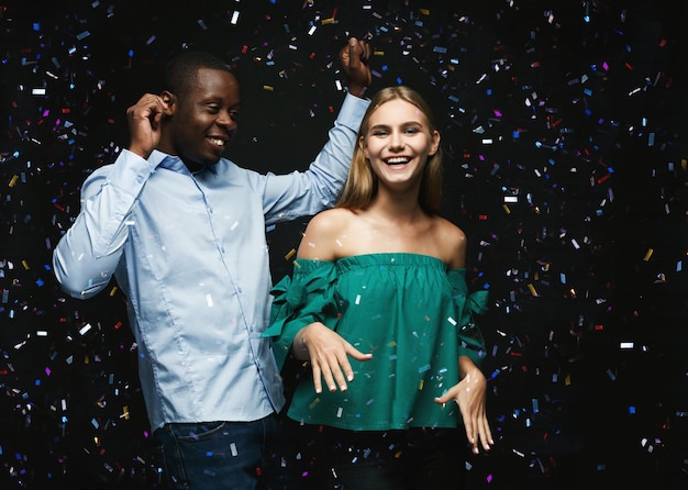 Birthday party at night club. Young couple, black man and smiling blonde girl feeling happy, dancing and celebrating, showered with confetti. Classy nightlife concept