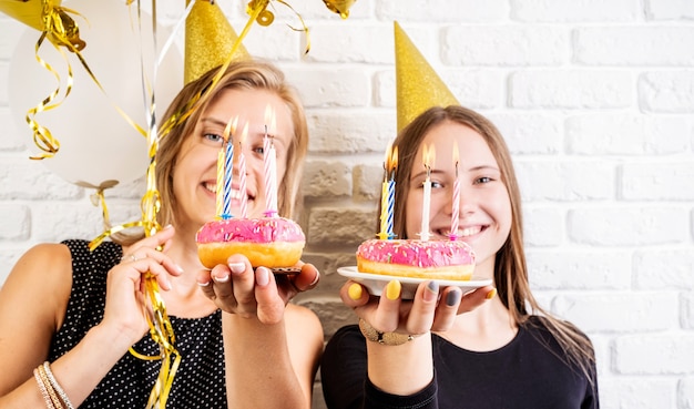 생일 파티. 흰색 벽돌 벽 배경 위에 촛불 도넛을 들고 생일을 축하하는 생일 모자에 두 웃는 젊은 여성 또는 자매