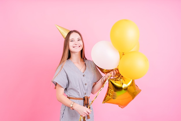 誕生日会。コピースペースでピンクの背景の上の誕生日パーティーを祝う風船と大きなギフトボックスを保持している誕生日の帽子の若い女性