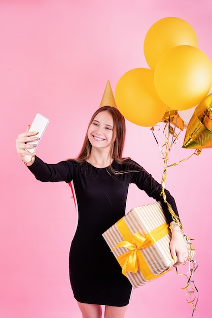 Birthday party. Young woman in a birthday hat holding balloons and big gift box celebrating birthday party taking selfie over pink background