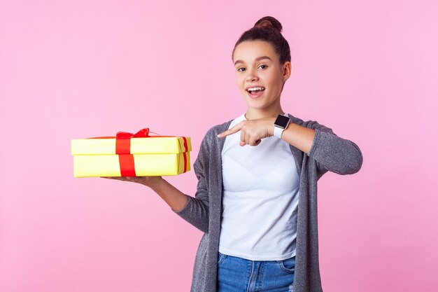 Birthday surprise present Portrait of happy teenage brunette girl with bun hairstyle in casual clothes pointing at gift box and smiling joyfully at camera studio shot isolated on pink background