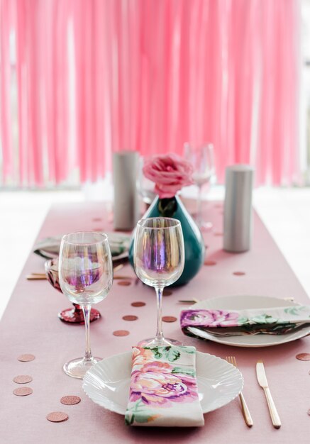 Birthday table setting in pink and colors with rose in vase. Streamers garland background. Baby shower or girl party.