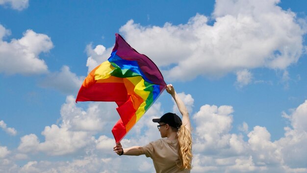 Bisexual girl lesbian woman transgender homosexsual holding in hand a rainbow LGBT gender identity flag on sky background with clouds on a sunny day and celebrating a gay parade in pride month