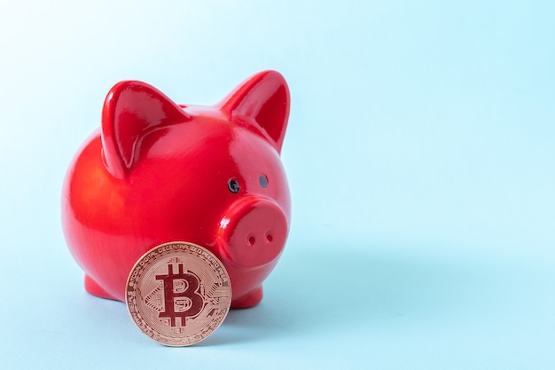 Bitcoin coin and red piggy bank on a blue background, close-up, copy space. Cryptocurrency saving concept. New virtual electronic and digital money