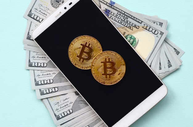 Bitcoins lies on a smartphone and hundred dollar bills on a light blue background