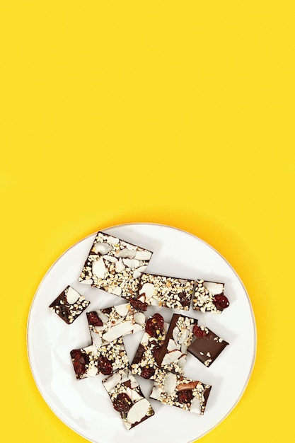 Bitten chocolate bar with nuts and  dried cranberry on white plate on yellow Sweet treat.