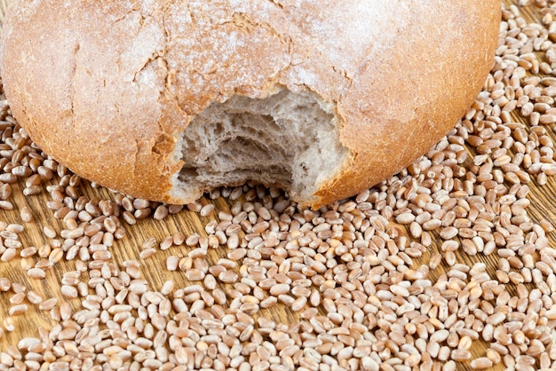 A bitten loaf of wheat bread lying on a wooden table along with whole grains