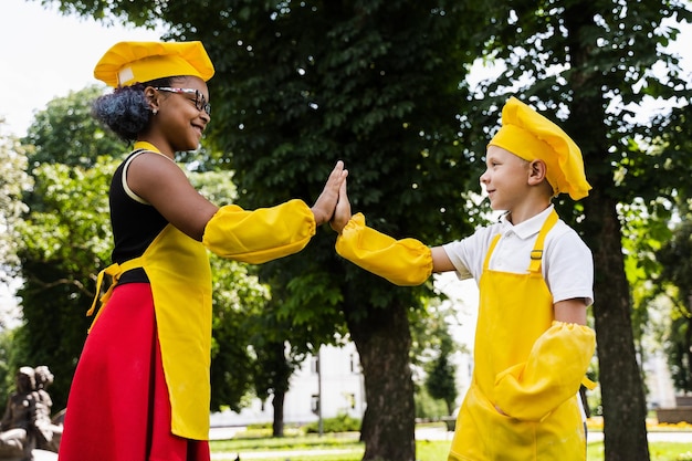 Black african and caucasian cook child in yellow chefs hat and apron clap each others hands and give five Black african sister cooking and caucasian brother smiling and having fun together