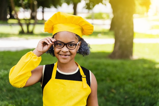 Black african child cook girl in chefs hat and yellow apron uniform touching hil glasses and smiling outdoor Creative advertising for cafe or restaurant