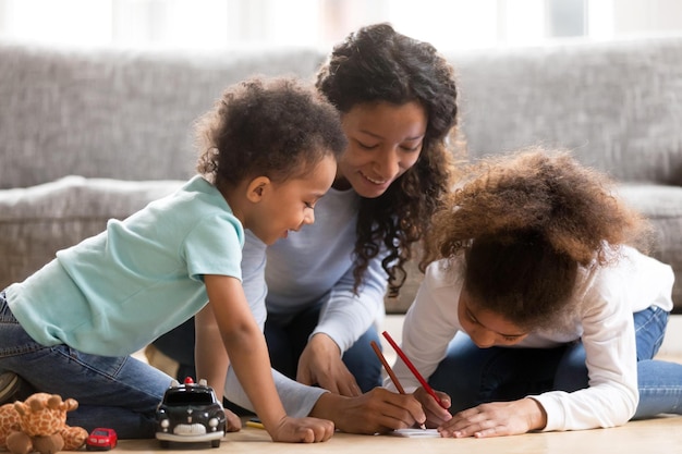 Black African loving mother help to their children draw Positive mom sitting with toddler son and preschool daughter on a wooden warm floor spend free time on weekend together in living room at home