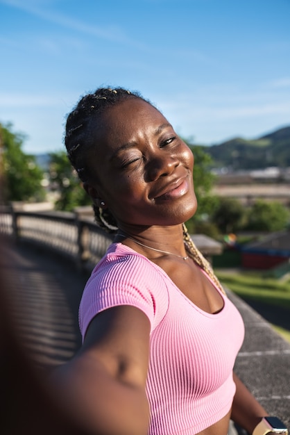 Black African woman taking a selfie holding the phone with her hands very happy and giving a kiss in a public park on a very sunny day and gray sky. Black woman in causal lifestyle clothing