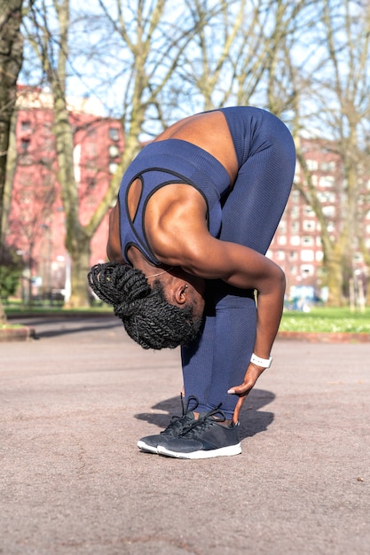 Photo black african woman warming up and doing back and leg stretches bending over holding her legs to start doing sports in a public park on a sunny day