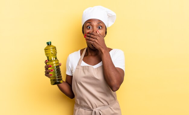 Black afro chef woman covering mouth with hands with a shocked, surprised expression, keeping a secret or saying oops. olive oil concept