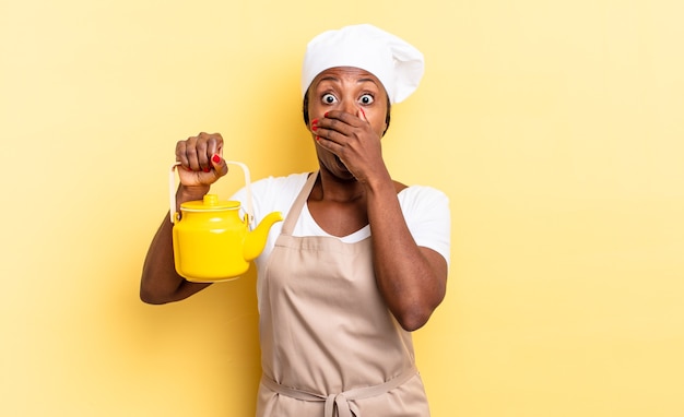 Donna chef afro nera che copre la bocca con le mani con un'espressione scioccata e sorpresa, mantenendo un segreto o dicendo oops. concetto di teiera