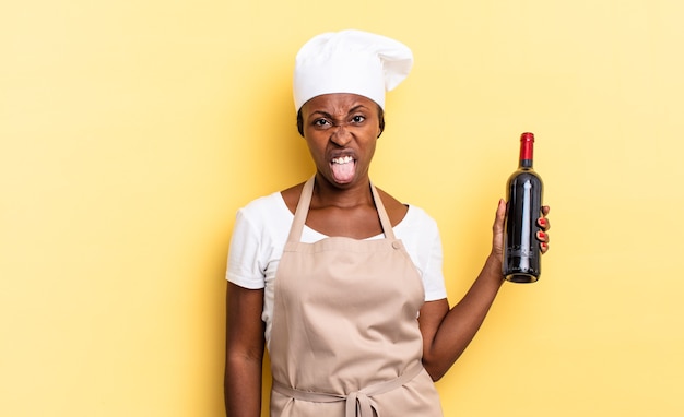 Black afro chef woman feeling disgusted and irritated, sticking tongue out, disliking something nasty and yucky. wine bottle concept