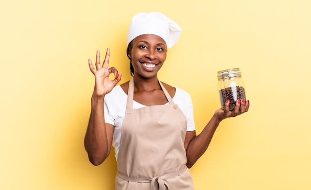 Черная афро-шеф-повар женщина чувствует себя счастливой, расслабленной и удовлетворенной, показывая одобрение с нормальным жестом, улыбаясь. кофе в зернах концепция