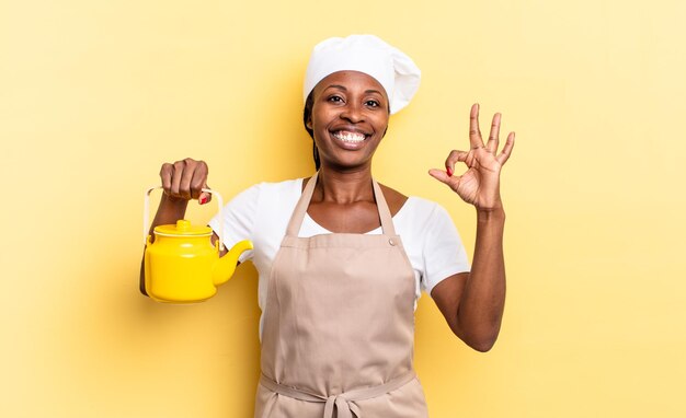 Black afro chef woman feeling happy, relaxed and satisfied, showing approval with okay gesture, smiling. teapot concept