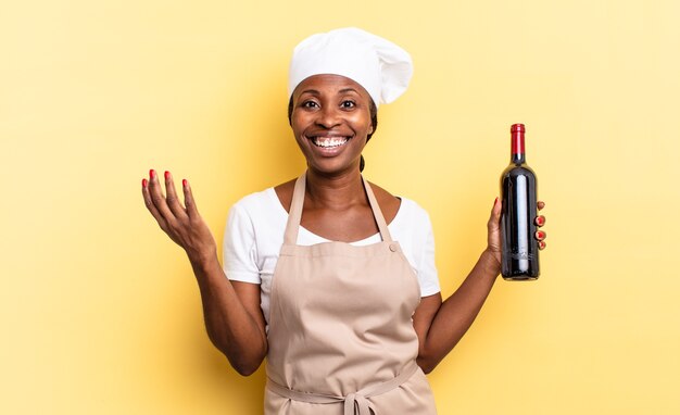 Черная афро-шеф-повар чувствует себя счастливой, удивленной и веселой, улыбается с позитивным настроем, реализует решение или идею. концепция бутылки вина