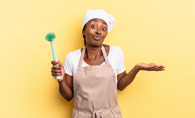 Foto donna chef afro nera che si sente perplessa e confusa, dubitando, ponderando o scegliendo diverse opzioni con un'espressione divertente. concetto di pulizia dei piatti