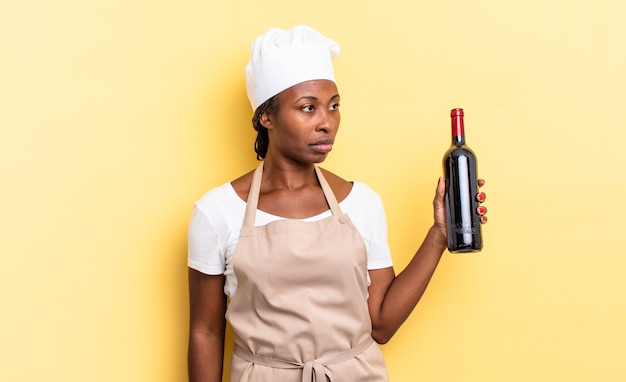 Donna chef afro nera che si sente triste, sconvolta o arrabbiata e guarda di lato con un atteggiamento negativo, accigliata in disaccordo. concetto di bottiglia di vino