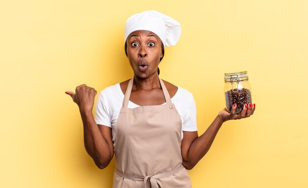 Black afro chef woman looking astonished in disbelief, pointing at object on the side and saying wow, unbelievable. coffee beans concept