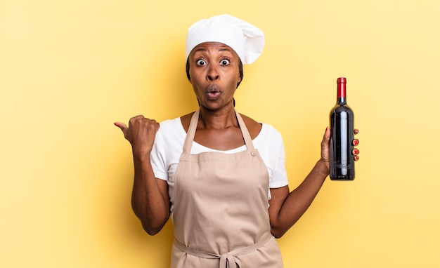 Donna afro chef nera che sembra stupita incredula, indicando un oggetto sul lato e dicendo wow, incredibile. concetto di bottiglia di vino