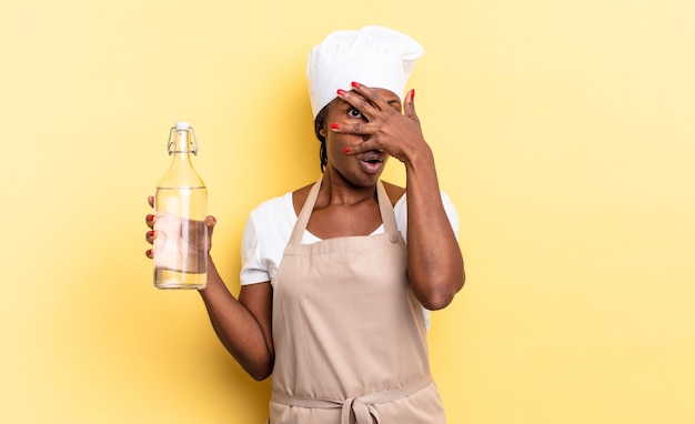 Black afro chef woman looking shocked, scared or terrified, covering face with hand and peeking between fingers holding a water bottle