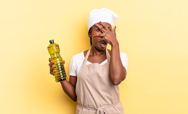 Black afro chef woman looking shocked, scared or terrified, covering face with hand and peeking between fingers. olive oil concept