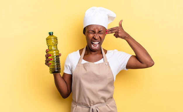 Donna chef afro nera che sembra infelice e stressata, gesto suicida che fa segno di pistola con la mano, indicando la testa. concetto di olio d'oliva