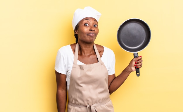 Black afro chef woman shrugging, feeling confused and uncertain, doubting with arms crossed and puzzled look