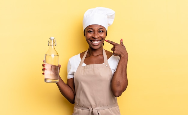 Black afro chef woman smiling confidently pointing to own broad smile, positive, relaxed, satisfied attitude holding a water bottle