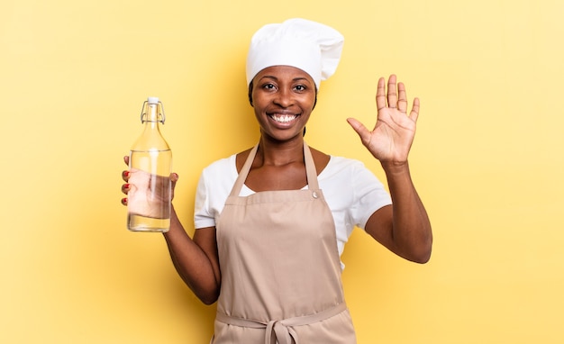 Черная афро-шеф-повар улыбается и выглядит дружелюбно, показывает номер пять или пятое с рукой вперед, считая, держа бутылку с водой