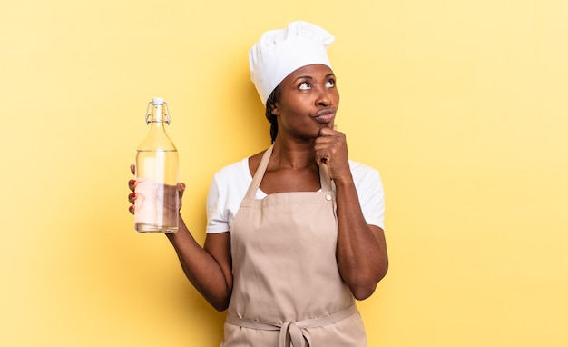 Black afro chef woman thinking, feeling doubtful and confused, with different options, wondering which decision to make holding a water bottle